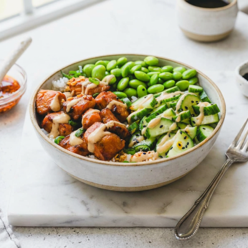 Vibrant high protein low calorie teriyaki salmon bowls with rice, edamame, cucumber, and a creamy miso-ginger yogurt drizzle.