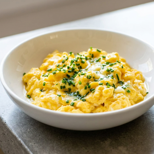 Fluffy high protein scrambled eggs, generously garnished with fresh green chives and melted butter on a white plate.
