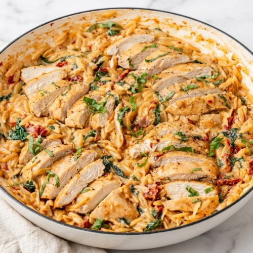 A large cast iron skillet filled with creamy tuscan chicken orzo, showing tender chicken pieces, wilted spinach, and sun-dried tomatoes in a rich cream sauce.