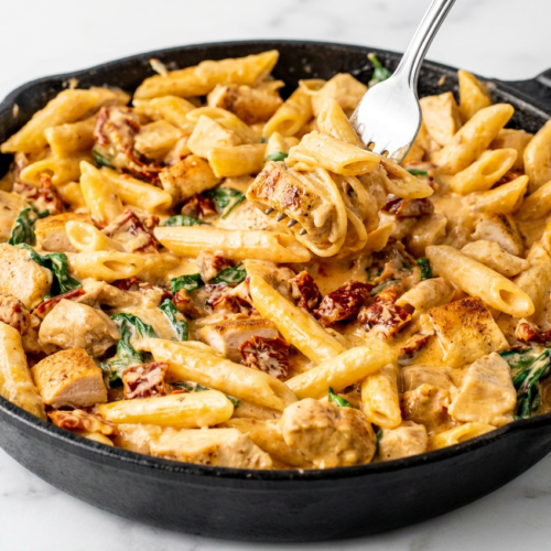 A white bowl of creamy tuscan chicken pasta, with tender chicken pieces, wilted spinach, and sun-dried tomatoes in a rich, savory sauce.