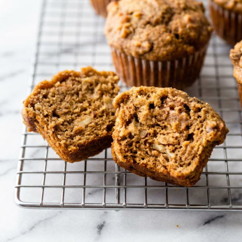 A stack of three golden brown high protein banana muffins, with one cut in half to show the moist interior, next to a fresh banana.