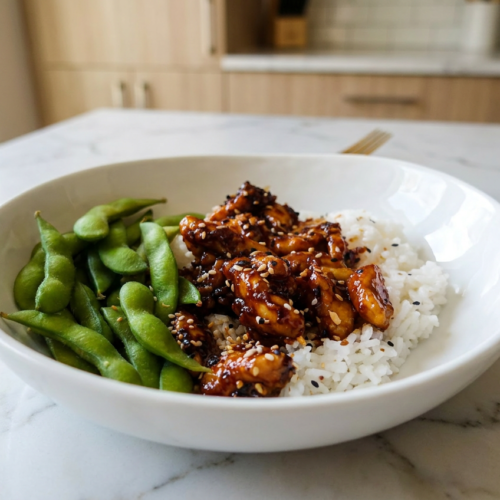 A serving of high protein mongolian chicken in a dark bowl, coated in a glossy soy-ginger sauce and garnished with sliced green onions and sesame seeds.