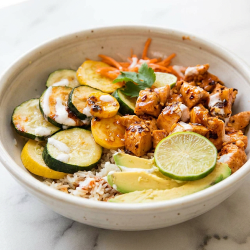 A close-up of hot honey chicken and rice bowls, topped with caramelized chicken, sliced avocado, and fresh zucchini, all drizzled with a glossy sauce.