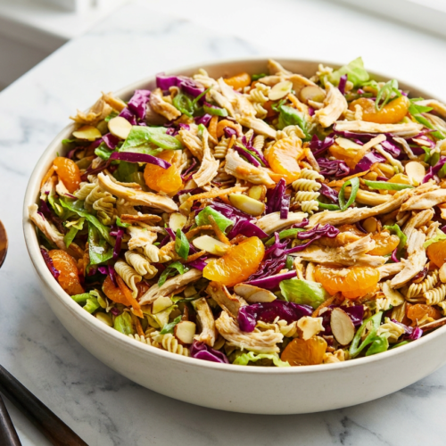 A large white bowl filled with mandarin chicken salad, featuring shredded chicken, romaine lettuce, bright orange mandarin segments, and a generous topping of crunchy toasted ramen noodles and sliced almonds.